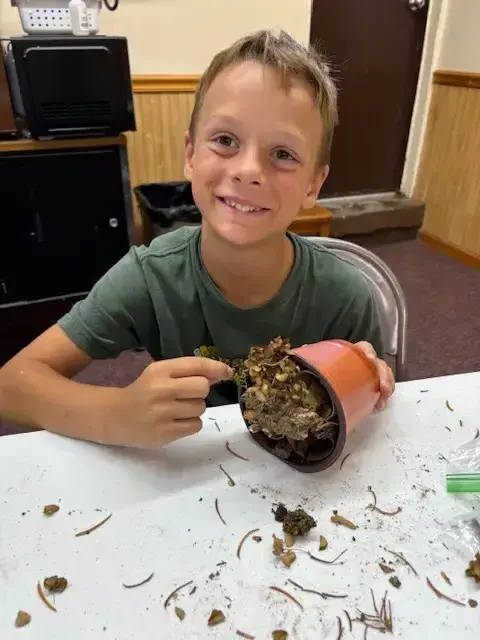 A smiling school-aged boy is at a white table, holding an orange plant pot that has spilled soil and dried plant material onto the table.