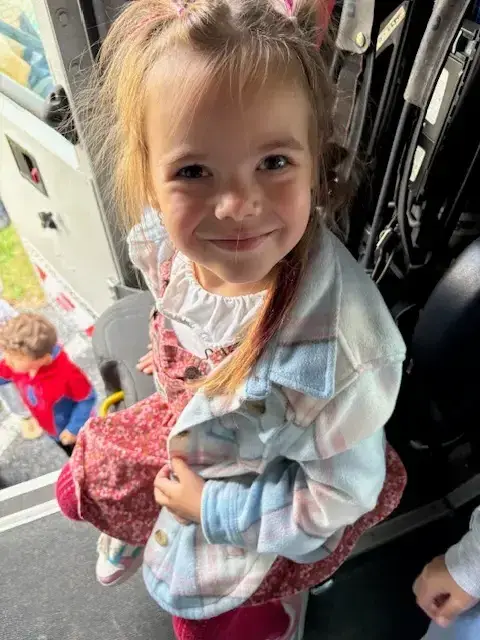 A smiling preschool-aged child wearing a plaid shirt over a floral dress looks up at the camera, with another child partially visible in the background.