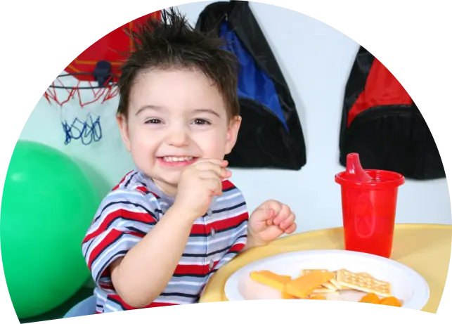 A smiling young boy in a striped shirt sits at a table with a plate of food and a red sippy cup, holding a piece of food to his mouth.