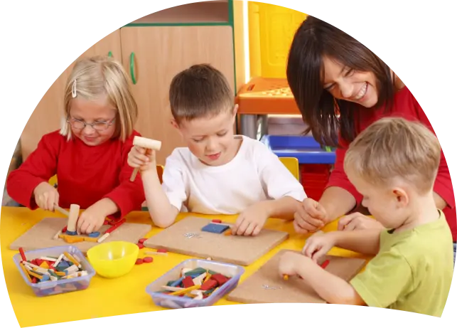 Three young children and an adult sit at a yellow table assembling small colorful shapes on cardboard pieces, with two divided trays of craft pieces and a yellow bowl nearby.