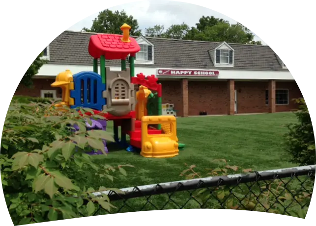 A colorful plastic play structure sits on a grassy lawn in front of a brick building with a sign reading HAPPY SCHOOL.
