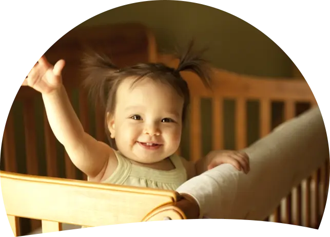 A baby with two small pigtails smiles and raises one arm while standing behind a wooden crib rail with a white blanket draped over it.