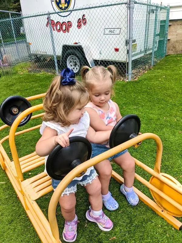 Two young girls sit together on a yellow playground glider with black steering wheels on green artificial turf, with a white trailer labeled 'TROOP 86' behind a chain-link fence.