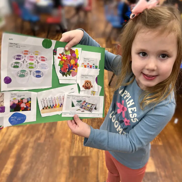 A young girl stands indoors holding a green display board covered with printed photos of art supplies and children's artwork.