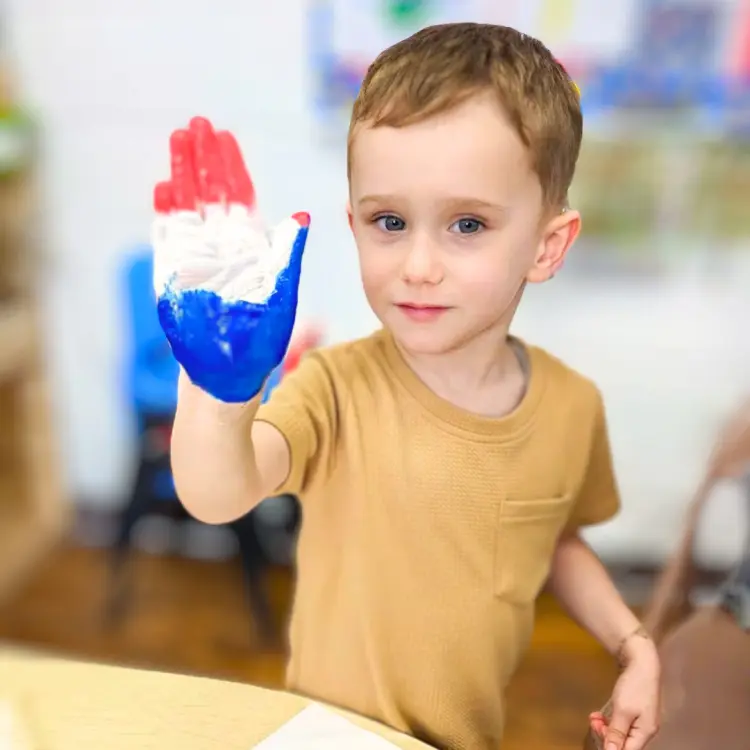 A young boy in a tan shirt holds up a hand painted with red, white, and blue paint.