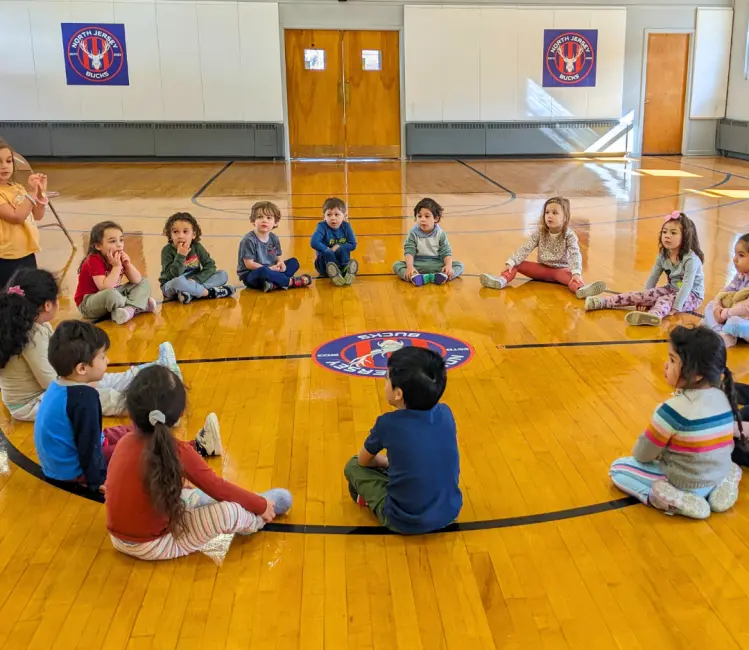 A group of children sit in a circle on a polished wooden gym floor while one child sits in the center facing away from the camera.