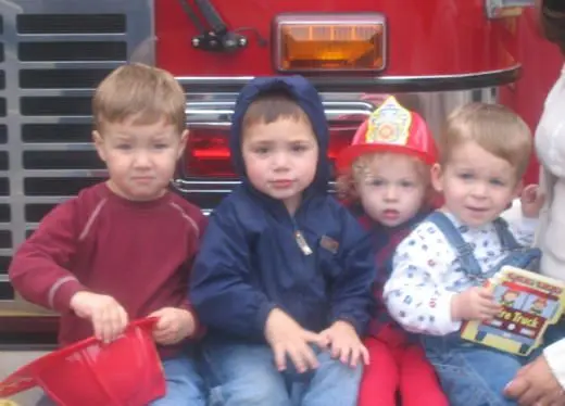 Four young children sit together in front of a red fire truck, with one child holding a red plastic container and another holding a book.