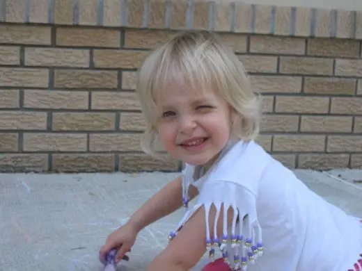 A smiling child with blonde hair sits on concrete in front of a brick wall, wearing a white top with beaded fringe and holding a purple marker.