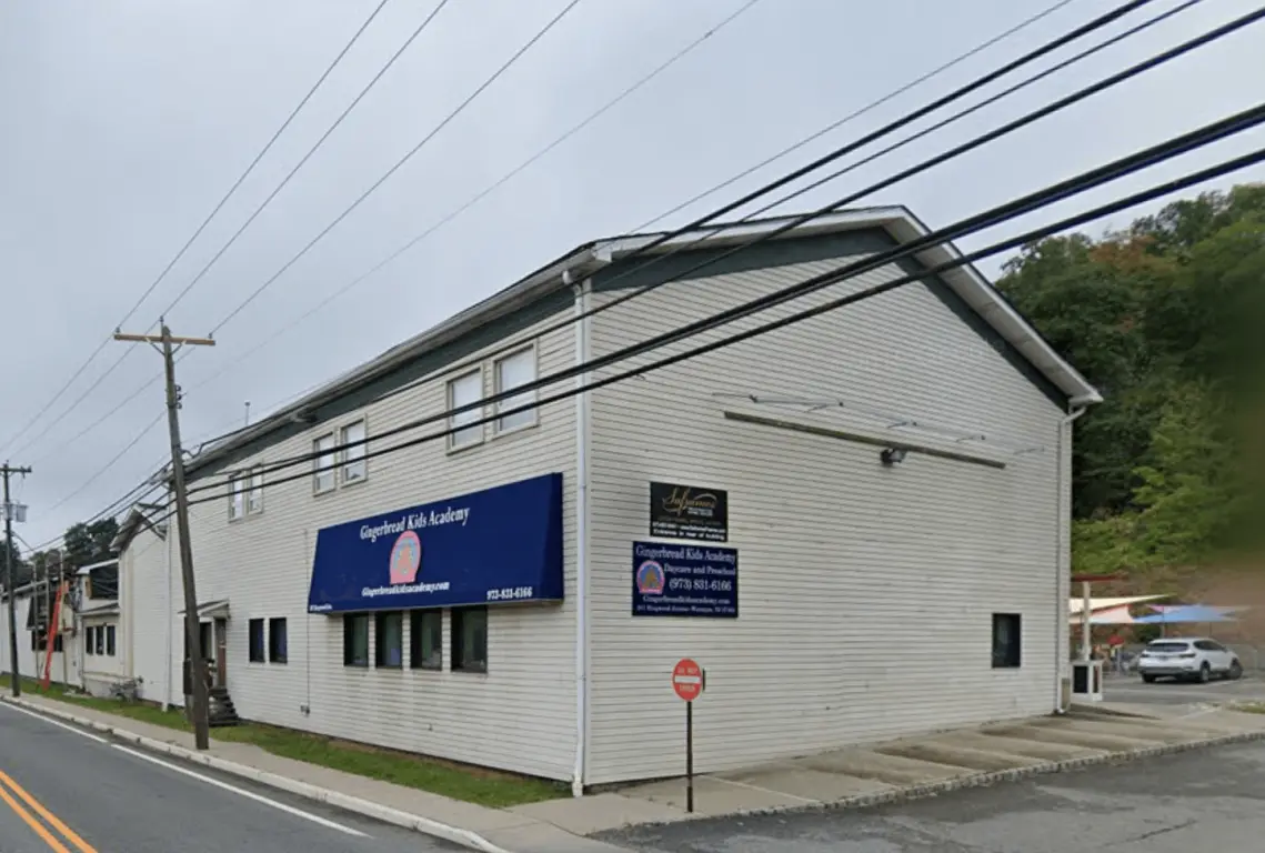 A two-story white building with a blue awning reading "Gingerbread Kids Academy" sits beside a paved road under an overcast sky.