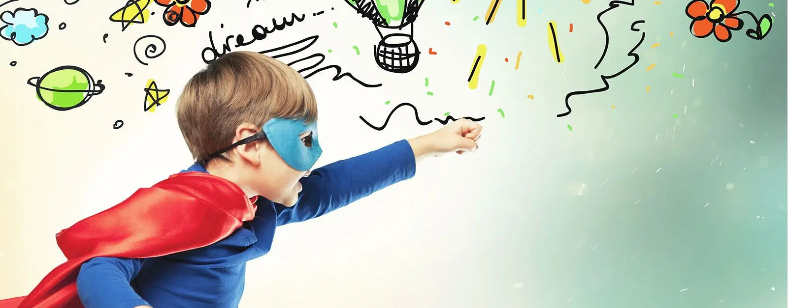 A young boy in a blue shirt, blue eye mask, and red cape poses with one arm extended forward against a wall covered in colorful childlike drawings.