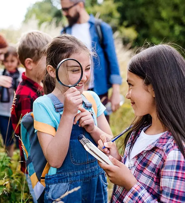 Two girls examine a notebook and a magnifying glass while three other people stand behind them in a grassy outdoor area.