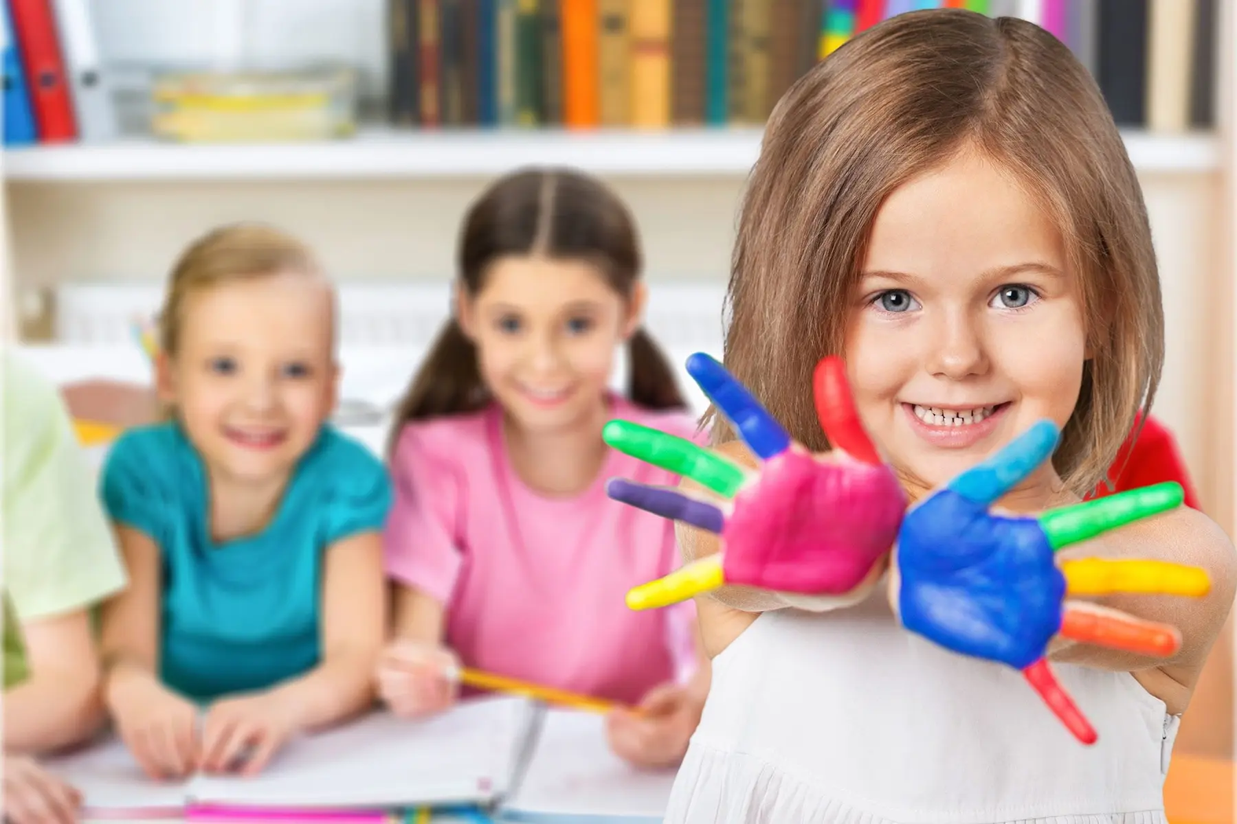 A girl in the foreground holds up two hands covered in bright finger paint while three other children sit at desks behind her.