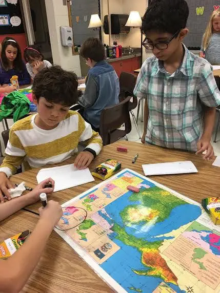 Two boys stand and sit at a table with a large map, markers, and snack boxes while other children work in the background.