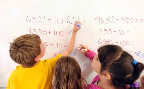 Three children stand at a whiteboard writing and solving arithmetic problems with markers.