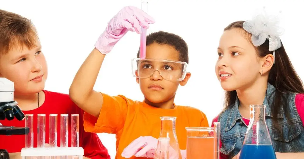 Three children look at a pink liquid in a test tube held up by a boy wearing safety goggles and pink gloves, with a microscope and glassware on the table.
