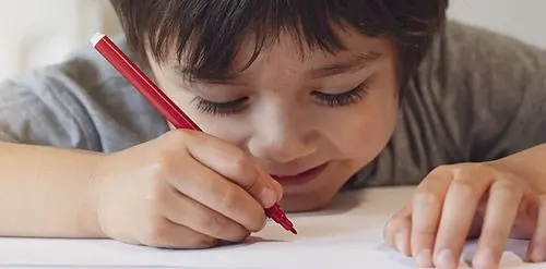 A young boy in a gray shirt holds a red pencil and draws on white paper while resting his other hand on the sheet.