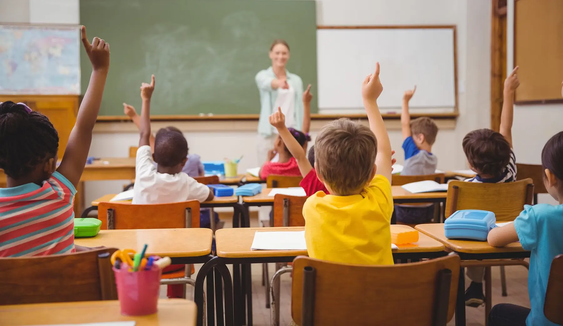 Several children sit at wooden desks with their backs to the camera, raising their hands toward a teacher standing near a green chalkboard.