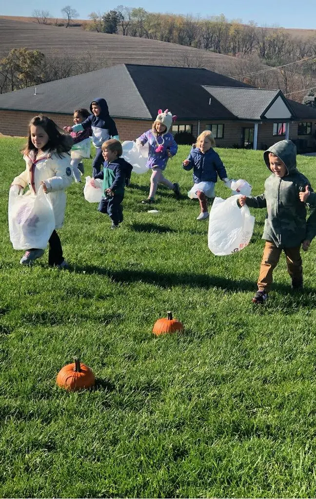 Seven children in coats holding plastic bags run across a grassy yard with pumpkins on the ground.