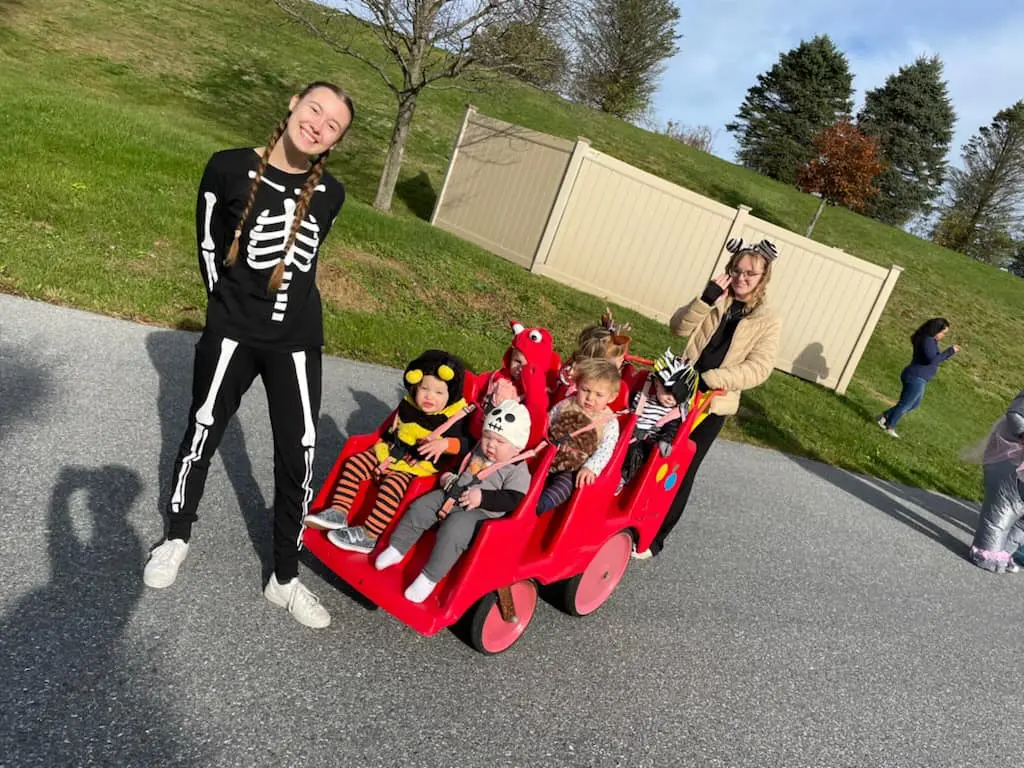 A person in a skeleton costume stands beside a red wagon with five children wearing costumes and one adult pushing the wagon on a paved path.