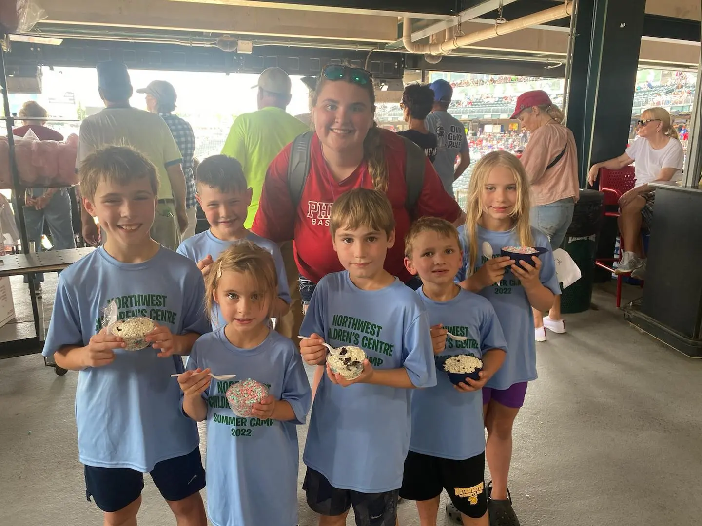 Group of children in matching blue shirts holding food items with an adult behind them at a stadium-like venue.