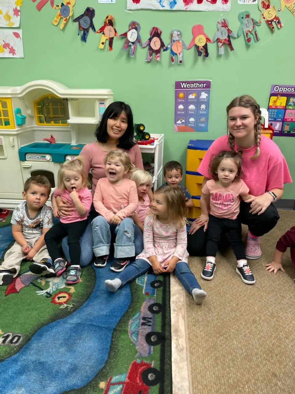 Two adults and six toddlers sitting on a colorful rug in a classroom with educational posters and toys.