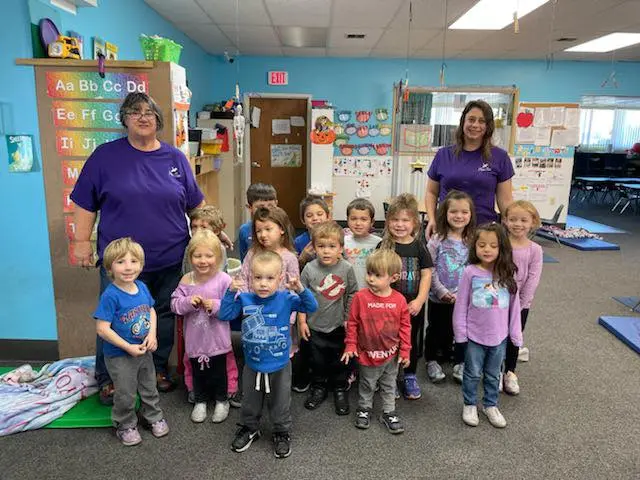 Group of preschoolers and two adults posing together in a classroom with alphabet letters on the wall.