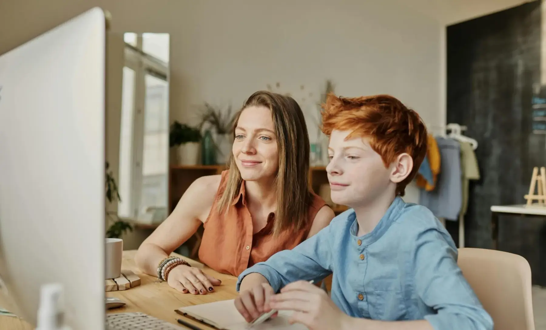 A woman and a boy sit at a desk looking at a computer monitor with the boy holding a notebook.