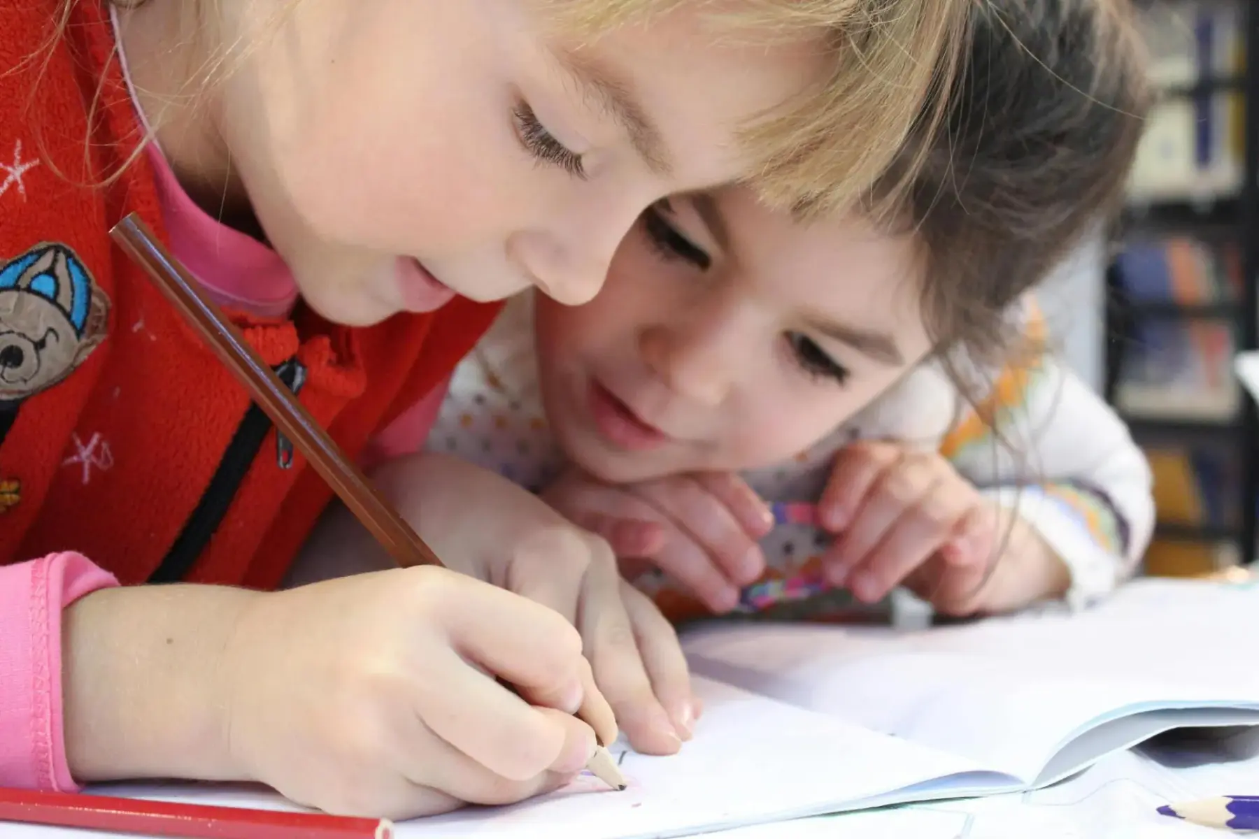 Two young children writing with pencils on a paper in an open book.