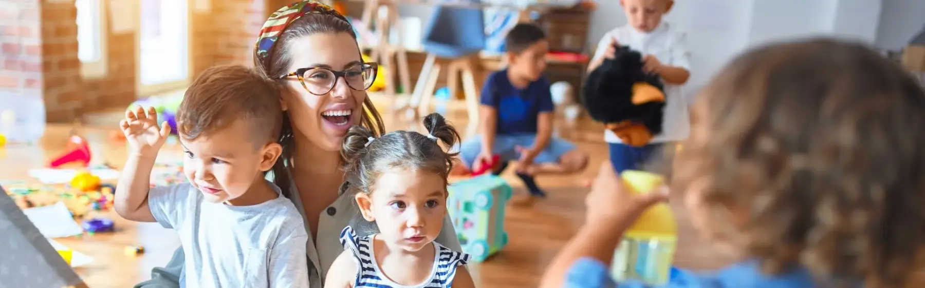 Adult holding two toddlers while other children play with toys in a room with wooden floors and scattered items.