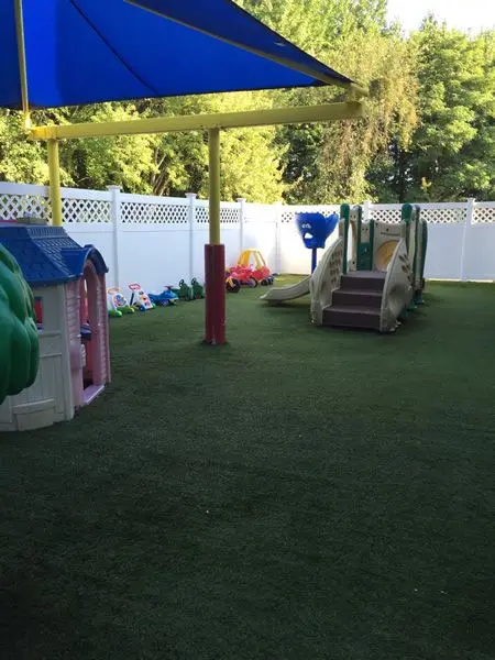 A fenced outdoor play area with artificial turf features a blue canopy, a small playhouse, a slide structure, and toy cars.