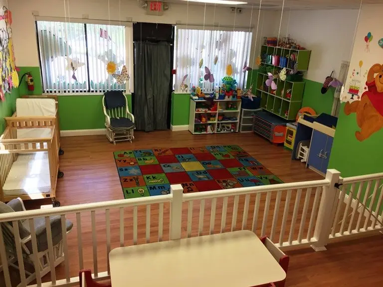 An empty childcare room with a wooden crib, glider chair, alphabet rug, low shelves of toys, and a white railing in the foreground.