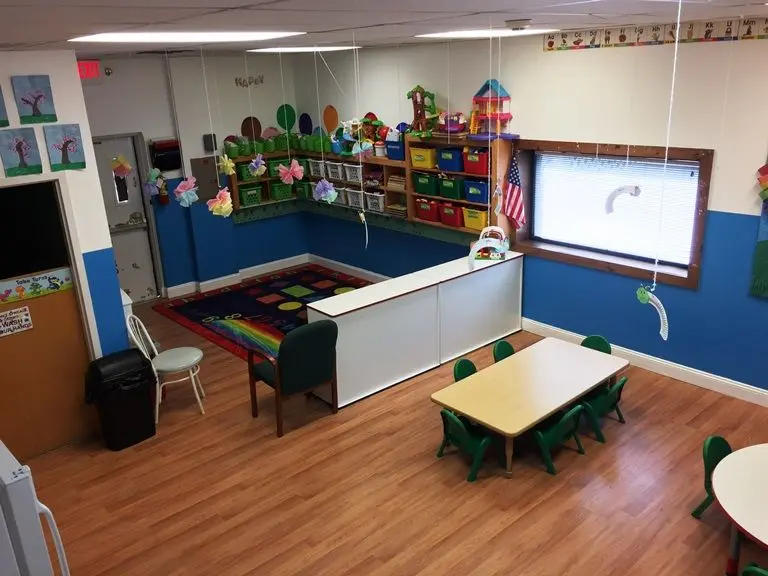 An empty preschool classroom with a colorful rug, low bookshelves, a whiteboard, a table with green chairs, and hanging paper decorations.