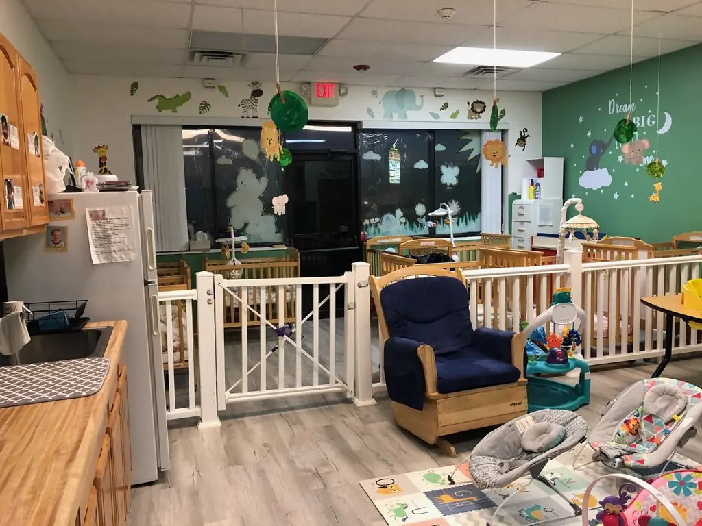 A childcare room with a white safety gate, wooden cribs, a blue armchair, a sink, a refrigerator, and animal wall decorations.