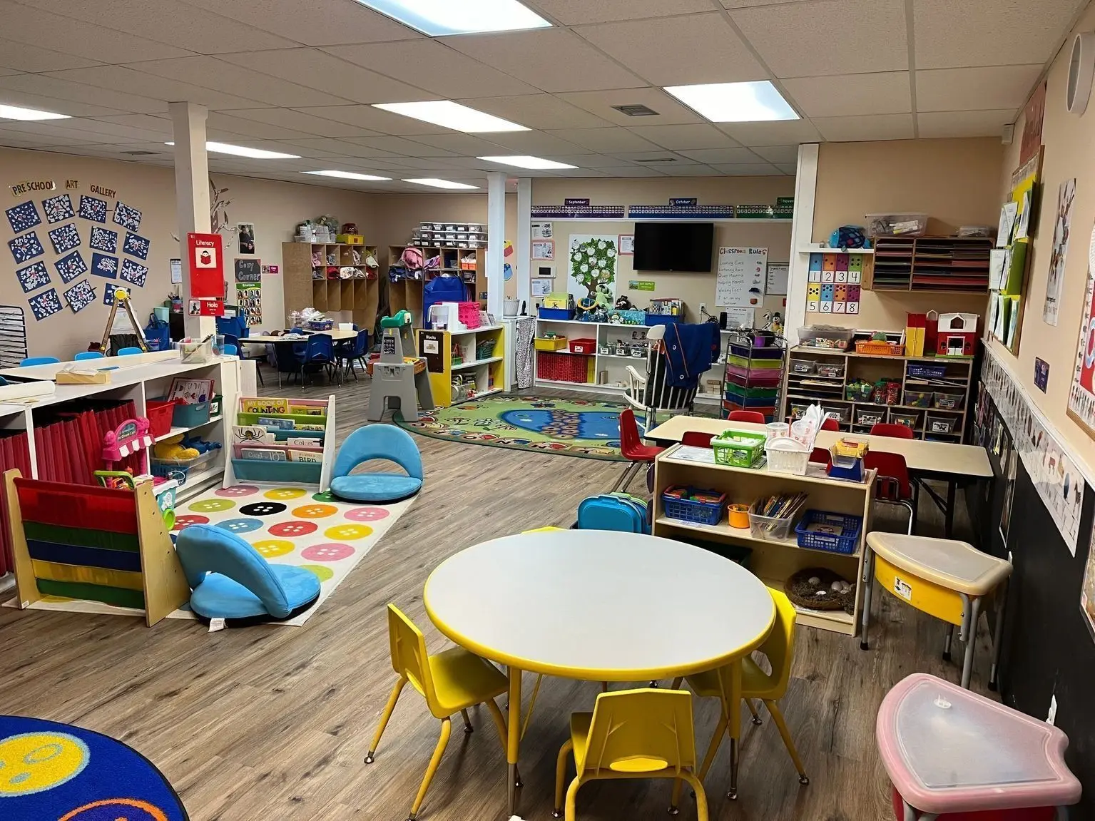 A brightly lit preschool classroom features small tables and chairs, colorful rugs, shelves filled with learning materials, and wall decorations.
