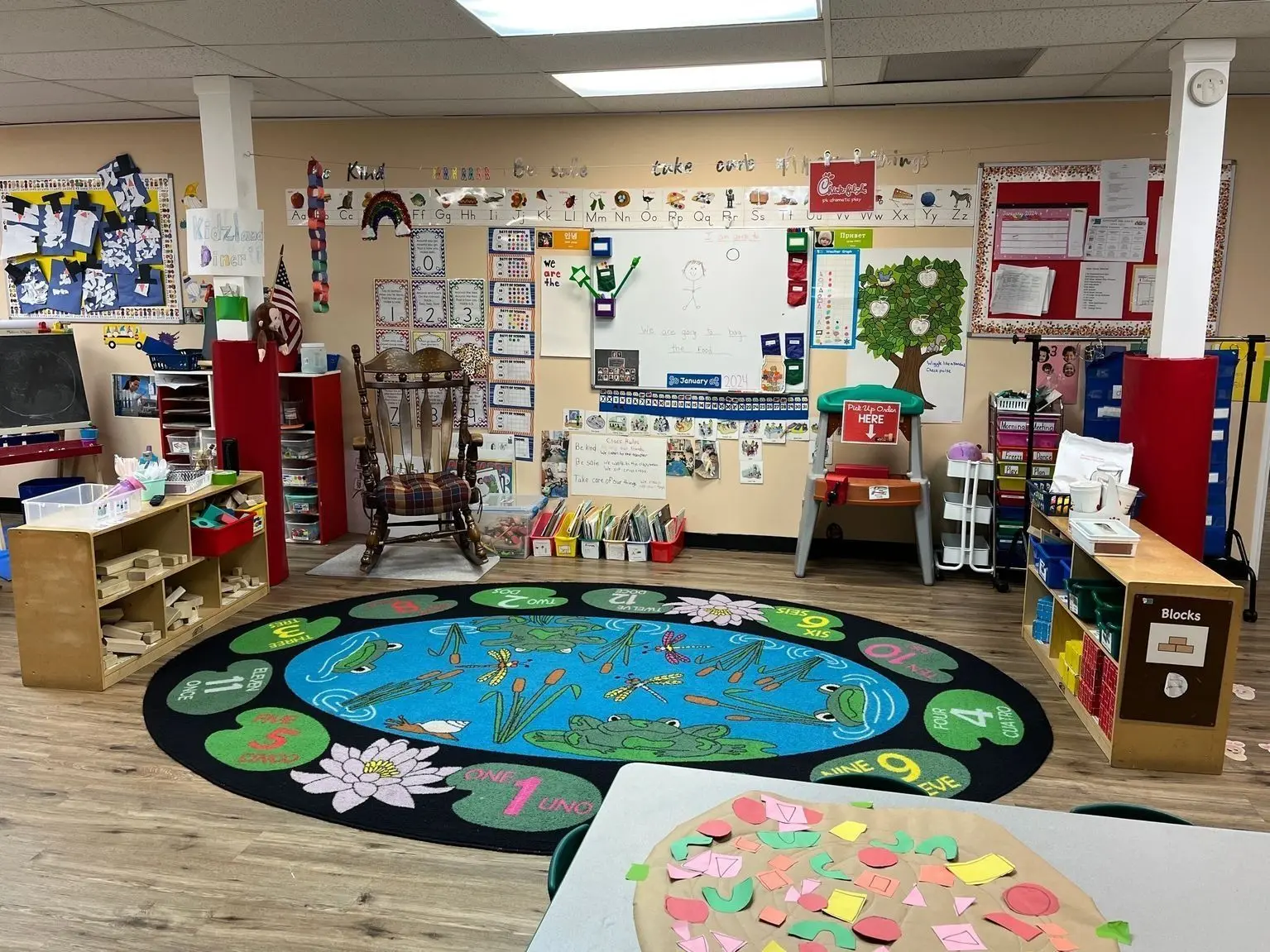 A brightly decorated preschool classroom features a large frog-themed rug, a rocking chair, various shelves with learning materials, a whiteboard, and a table with craft supplies.