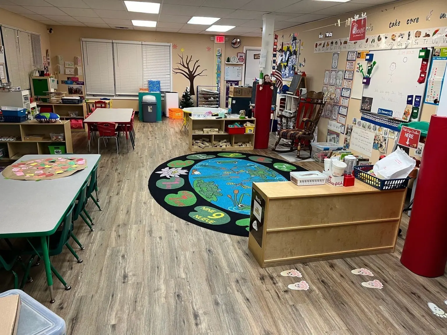A brightly lit classroom features small tables and chairs, a colorful frog-themed rug, shelves filled with learning materials, and a whiteboard on the wall.