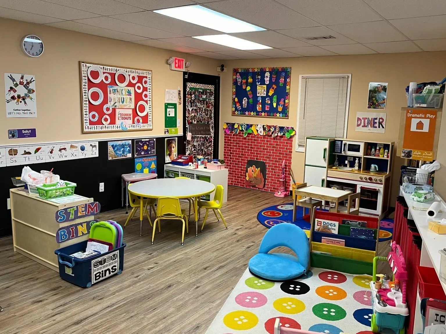 A colorful classroom features small yellow tables and chairs, a play kitchen, a dramatic play area, STEM bins, and various educational decorations on the walls.