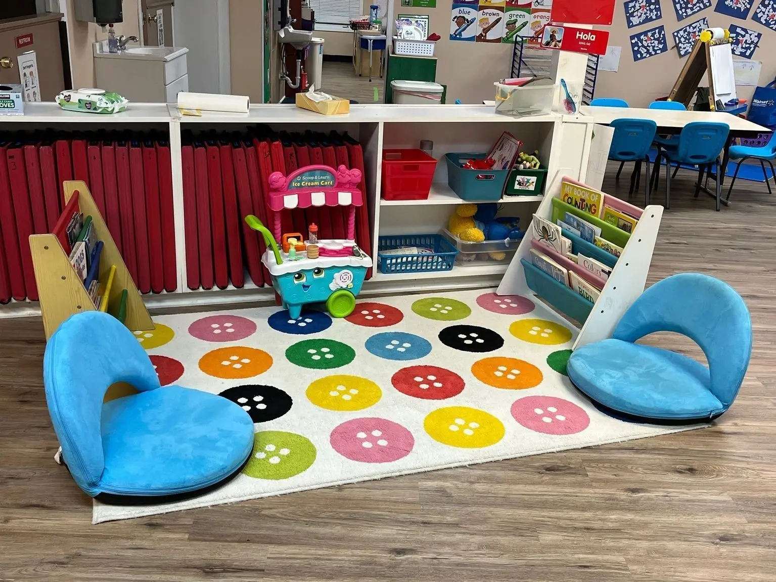 An empty classroom features two blue floor chairs on a colorful button rug, a play ice cream cart, two book shelves, and white storage units filled with toys and learning supplies.