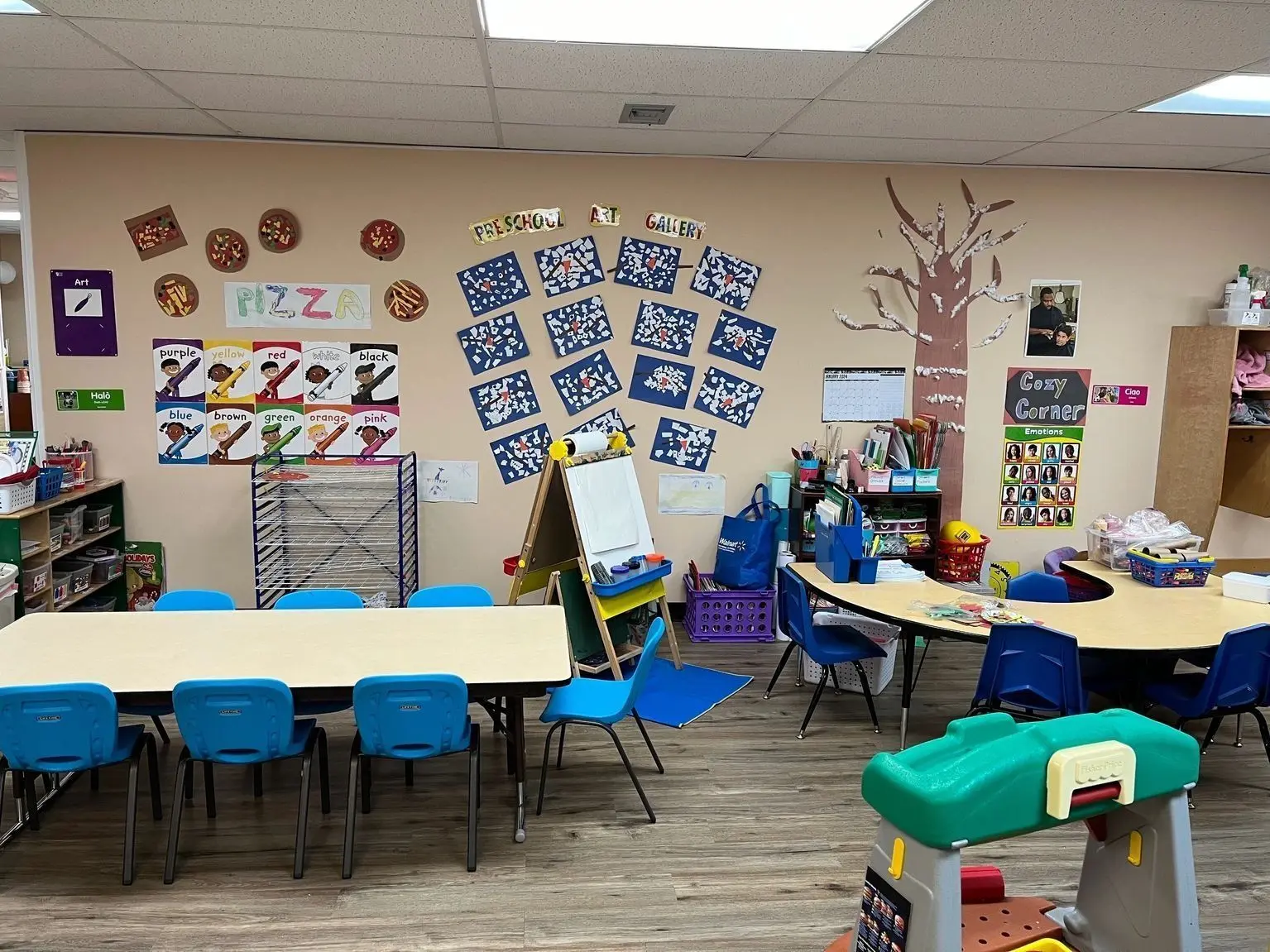 A brightly decorated preschool classroom features small tables with blue chairs, an easel, a drying rack, and various art projects and educational charts on the walls.