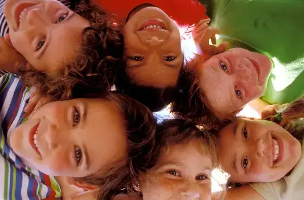 Six children lean inward in a tight circle, smiling down toward the camera against a bright sky.