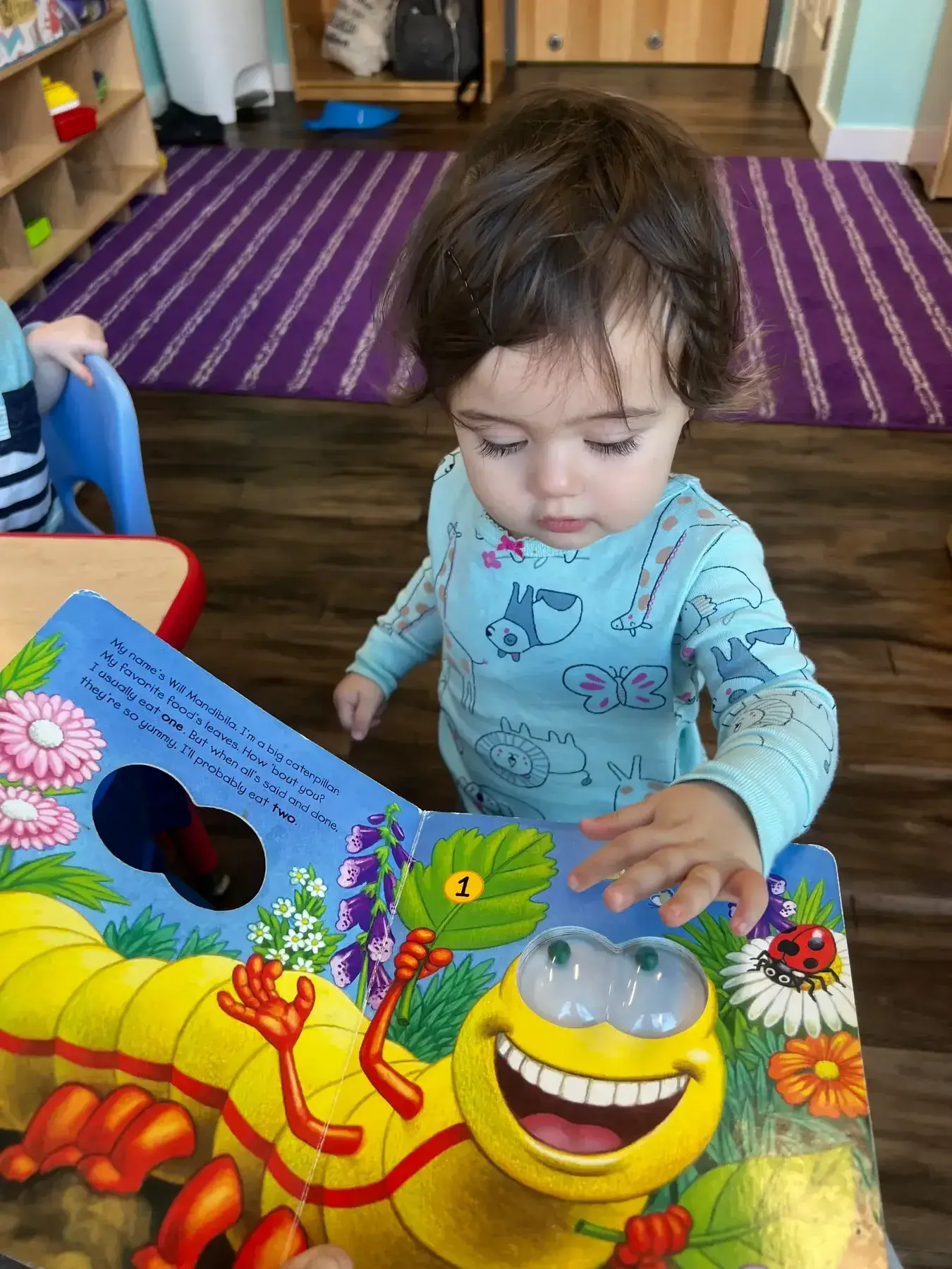 A young child in patterned pajamas looks down while touching a colorful board book featuring a cartoon caterpillar with googly eyes.